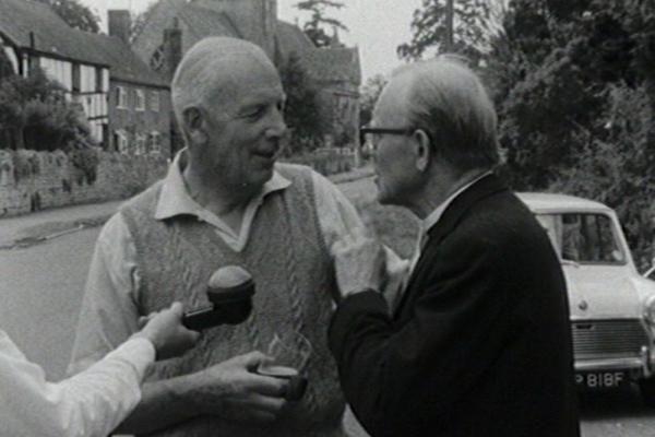 black and white image of a priest and fellow villager being interviewed. 